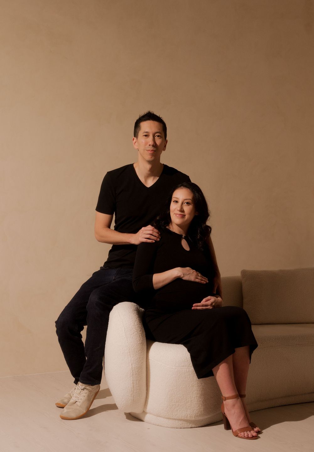 Couple in matching black outfits poses for maternity photo on white sculptural furniture against beige backdrop.