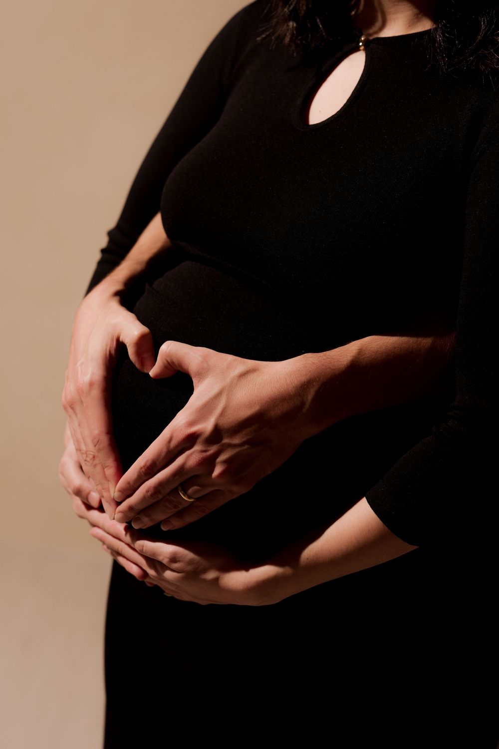 Pregnant woman in black dress with partner's hands forming heart shape over baby bump on beige background.