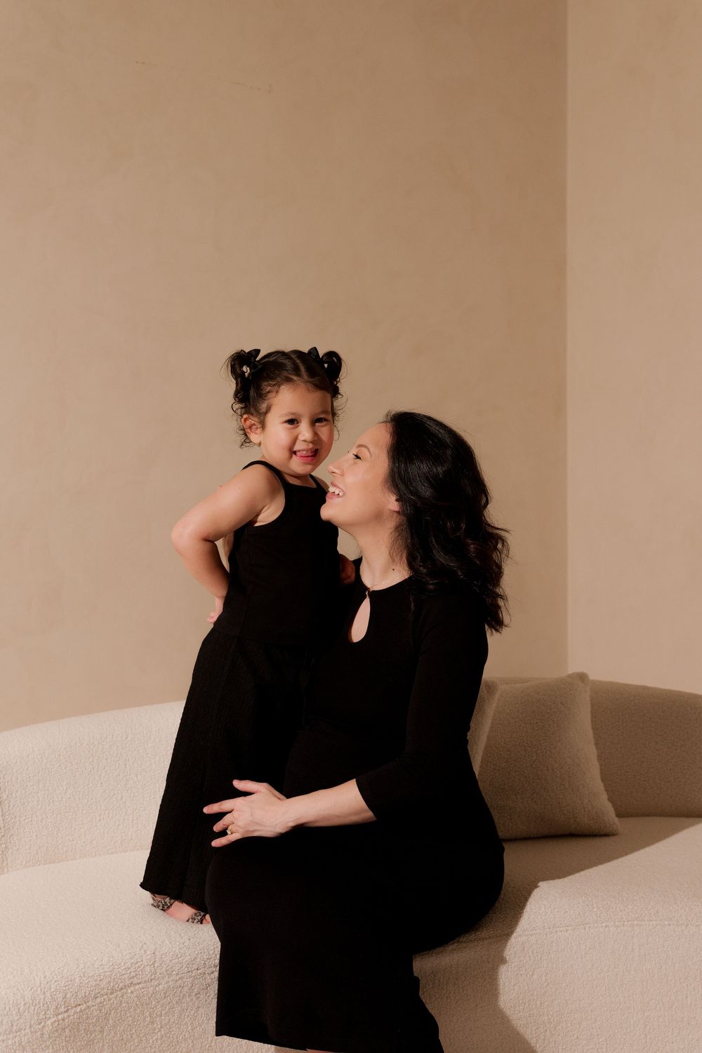 A mother and daughter in matching black dresses share a joyful moment on a cream sofa against a beige wall.
