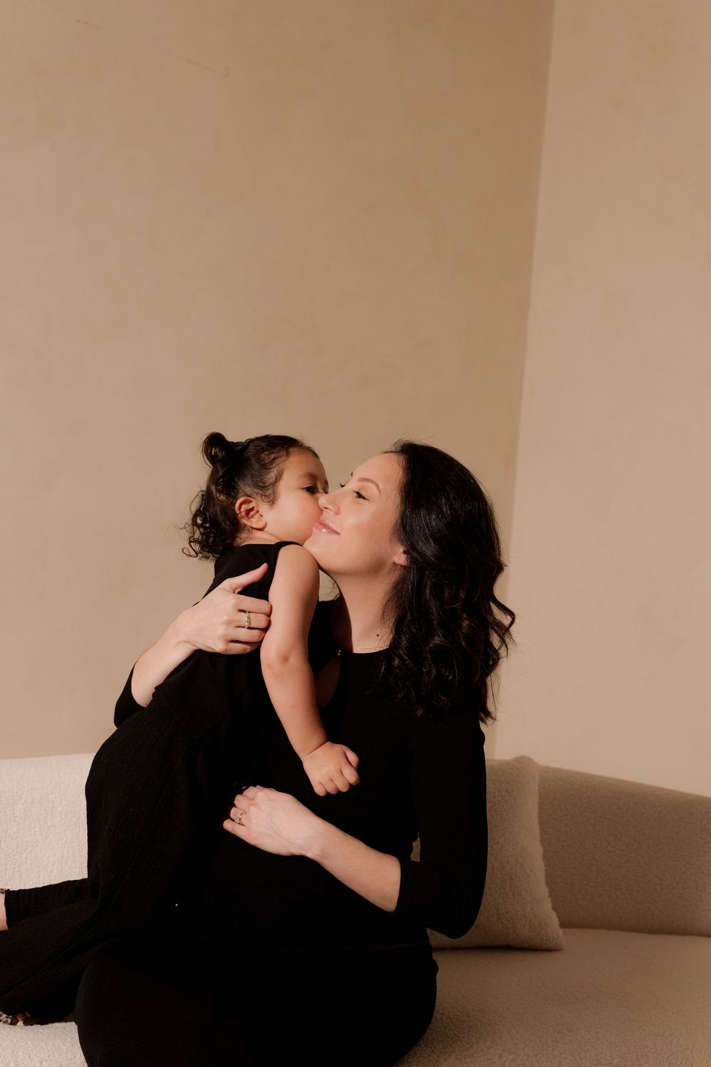 A mother in black holds her toddler who gives her a kiss on the cheek while sitting on a couch.