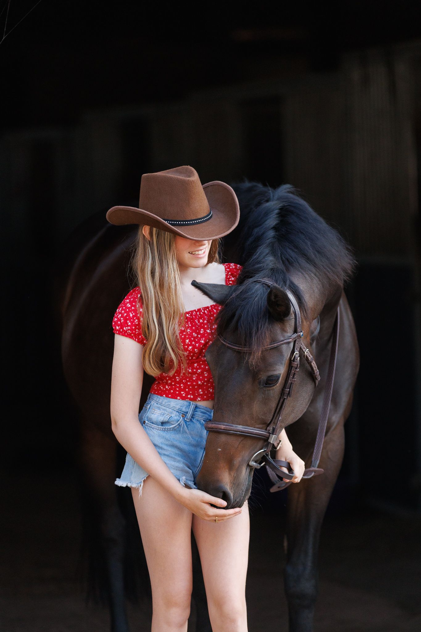 Young woman in a brown cowboy hat, red floral top and denim shorts stands affectionately beside a dark bay horse in a dim stable.