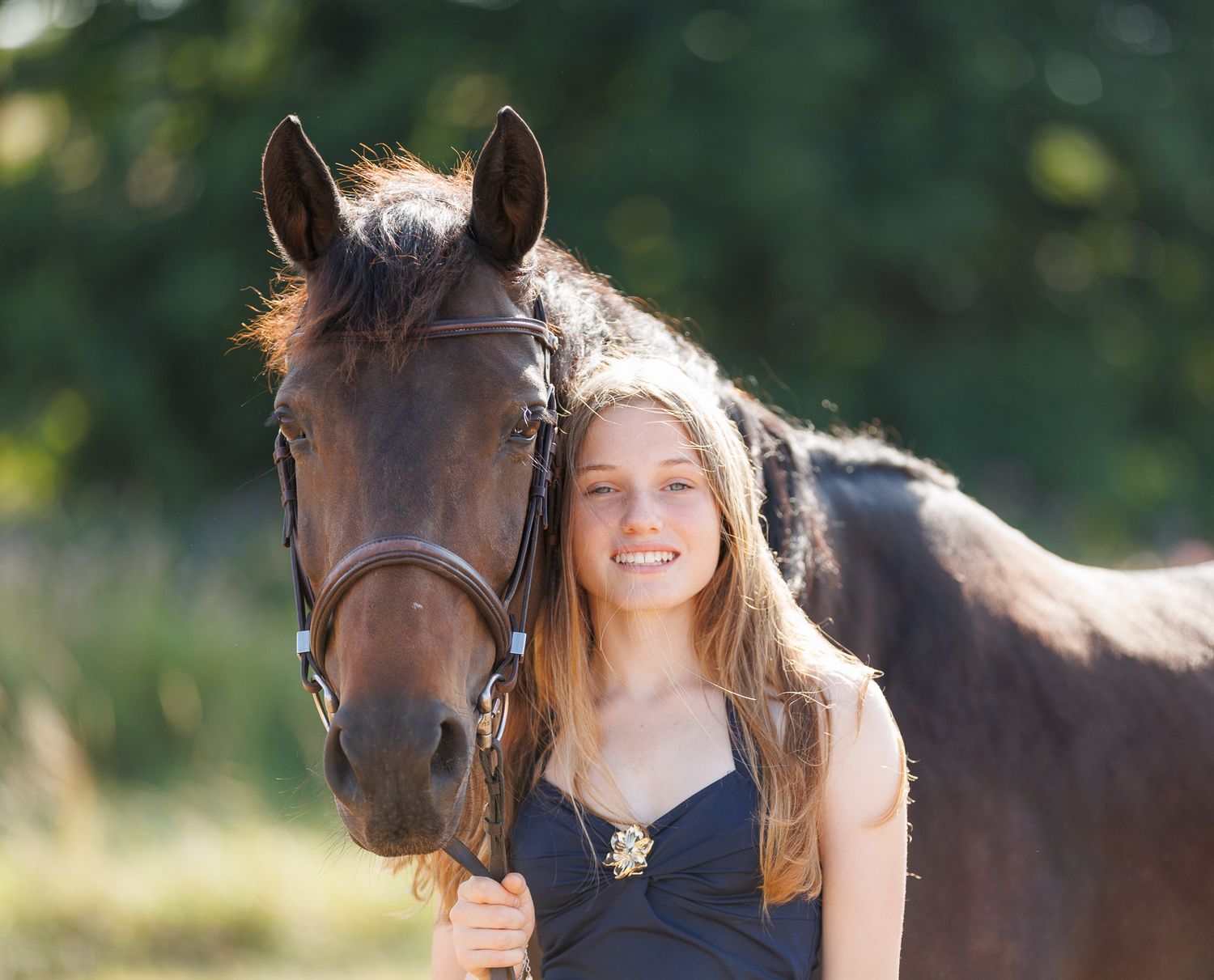Young woman in black top smiling while gently embracing a brown horse outdoors among lush green trees.