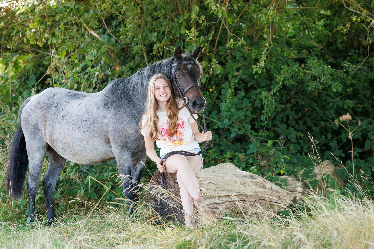Smiling girl with long hair sits on a log beside a gray pony wearing a halter, surrounded by green foliage and tall grass.
