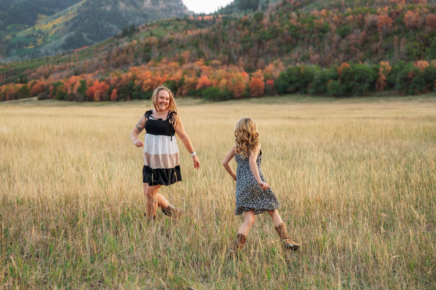 Two girls in dresses run through a golden meadow with colorful autumn mountain foliage in the background.