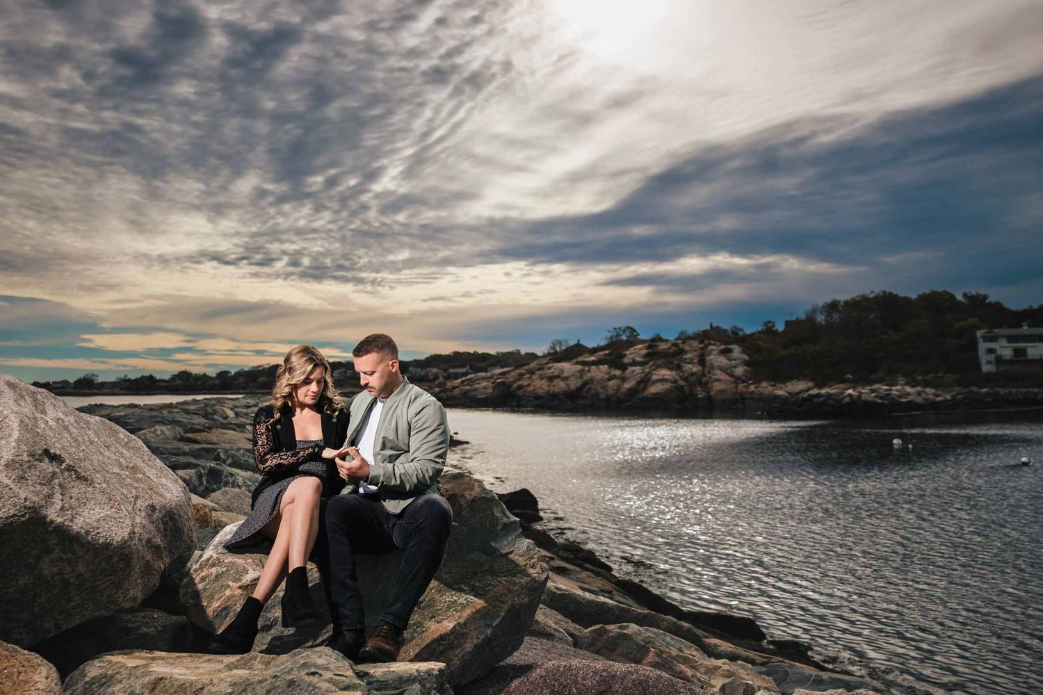 Couple sitting together on rocky breaker, gazing at each other with the water and sky behind them.  Engagement at Rockport Harbor, Rockport