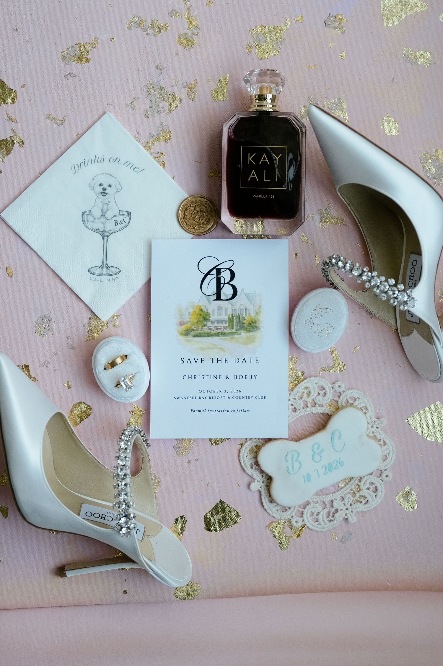 Wedding details flat lay with a save-the-date card, perfume, crystal bridal heels and a dog-bone cookie on a pink background.