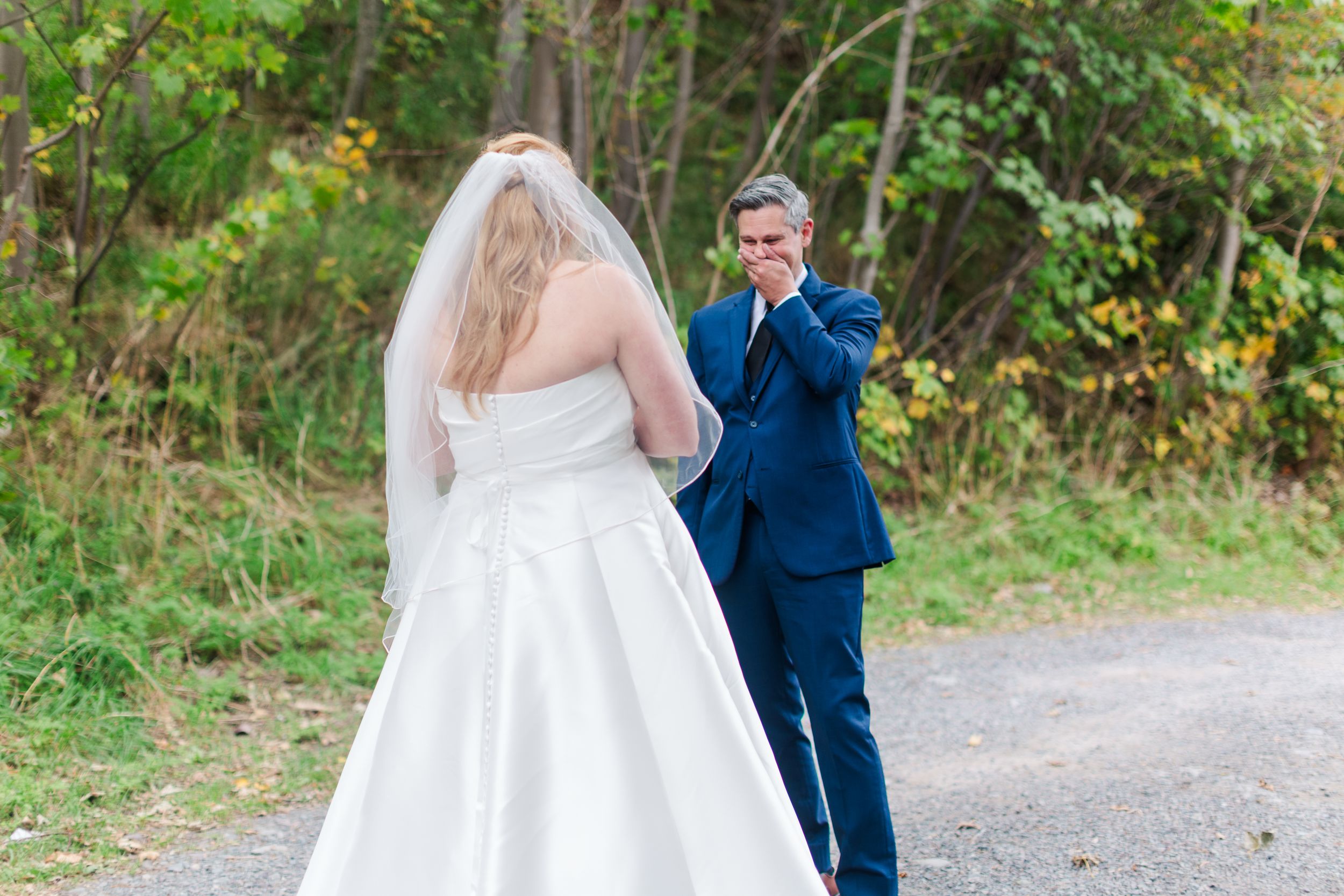 Bride in a white gown and veil faces a groom in a navy suit who covers his mouth, moved, during their first look on a wooded path.