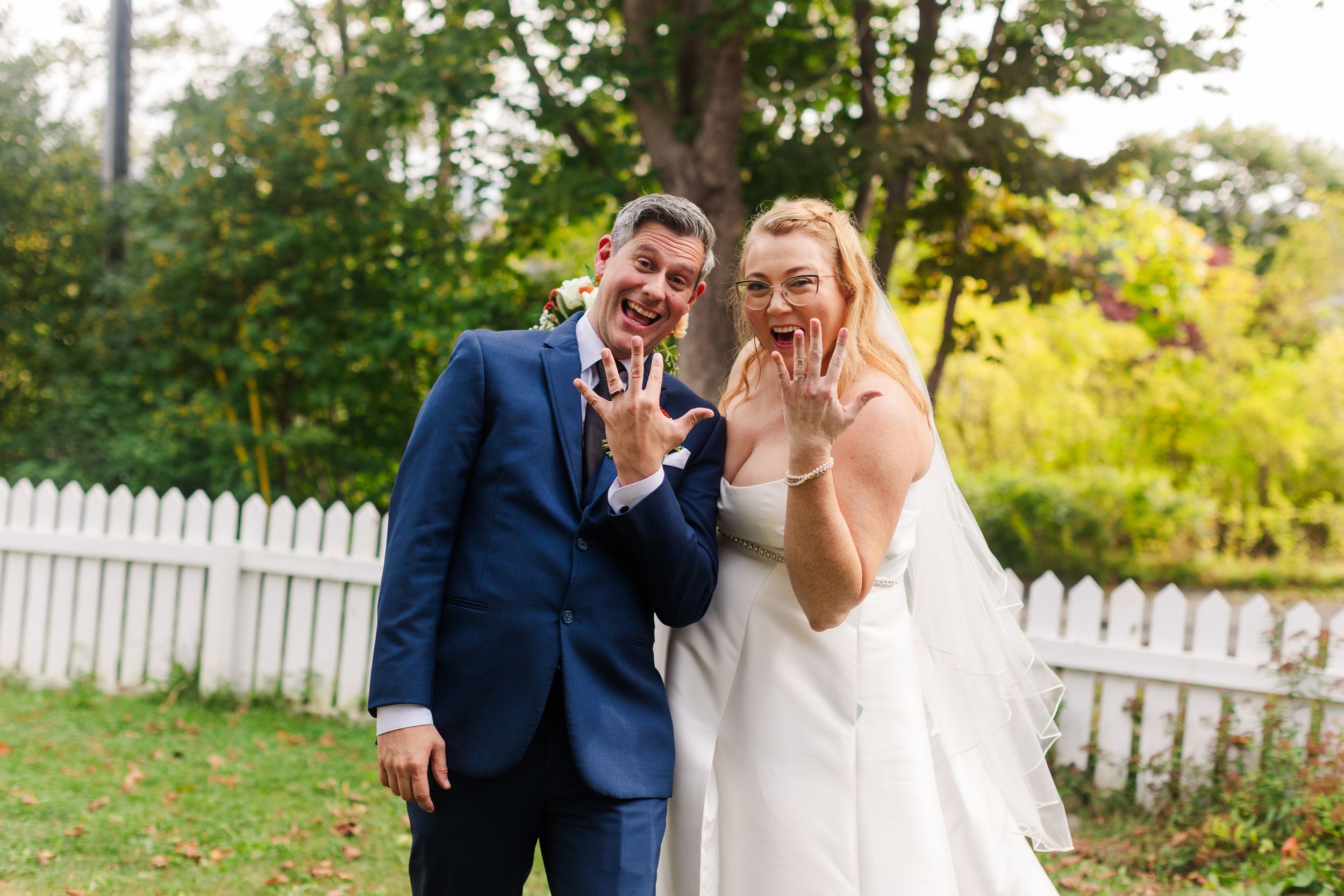 Smiling bride in a white gown and veil and groom in a navy suit showing off their wedding rings in a sunny garden with a white picket fence.