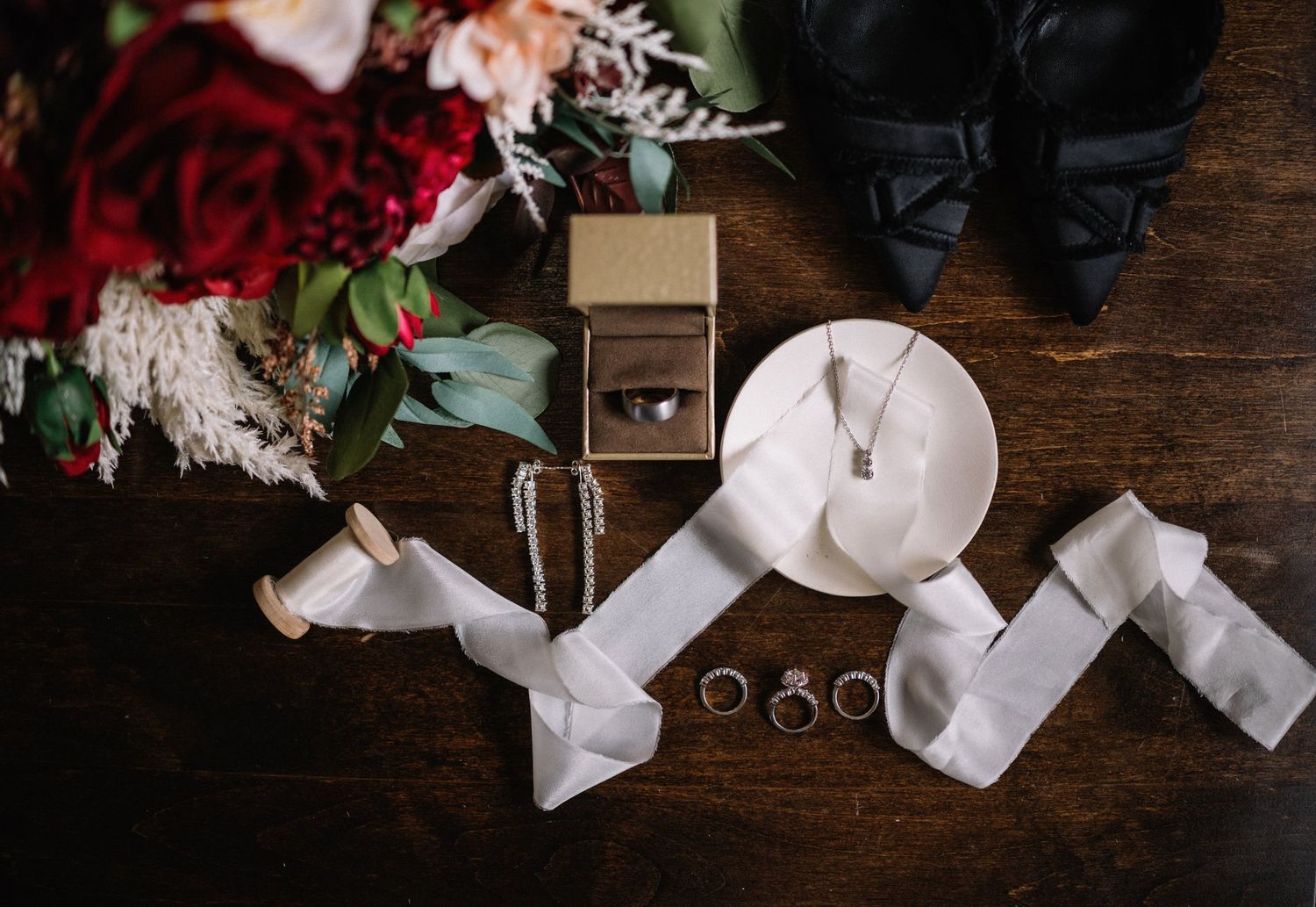 Winter wedding flat lay with red roses, black heels, ring box, custom wedding rings, earrings and ivory ribbon on dark wood