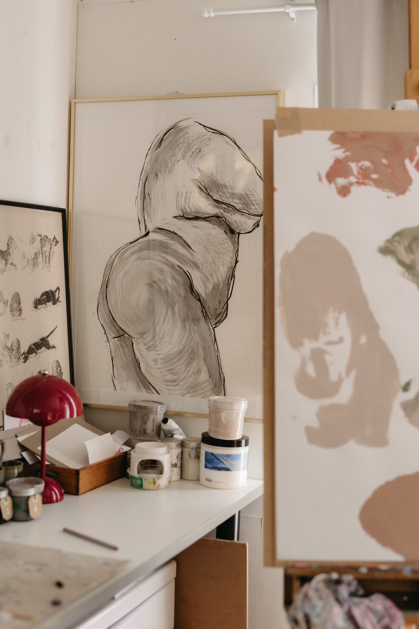 Artist studio with charcoal figure drawing on easel, paint supplies on white desk, and abstract canvas in foreground.
