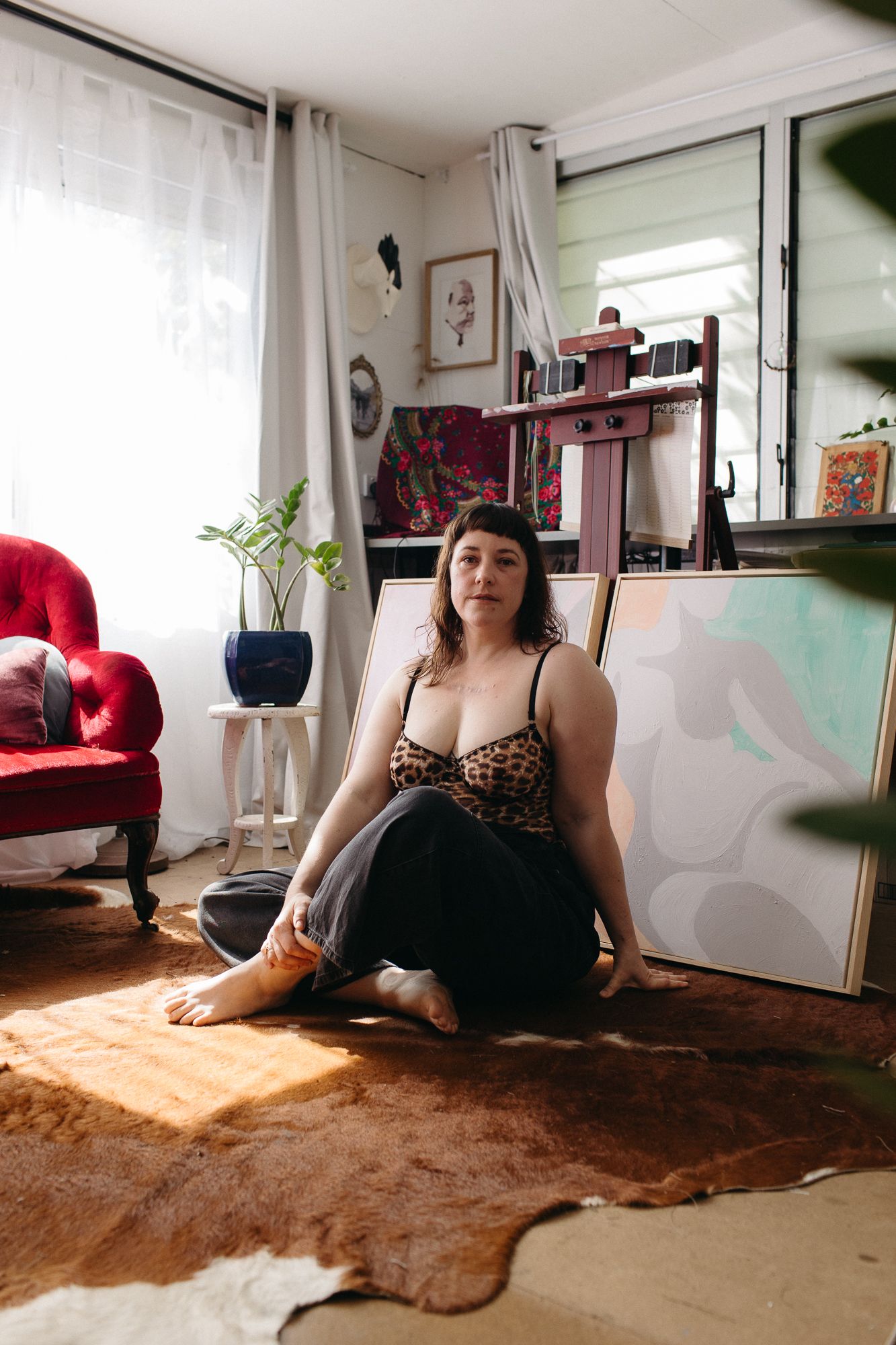 Artist sitting on a cowhide rug in her sunlit studio, surrounded by canvases and an easel, wearing a leopard print top.