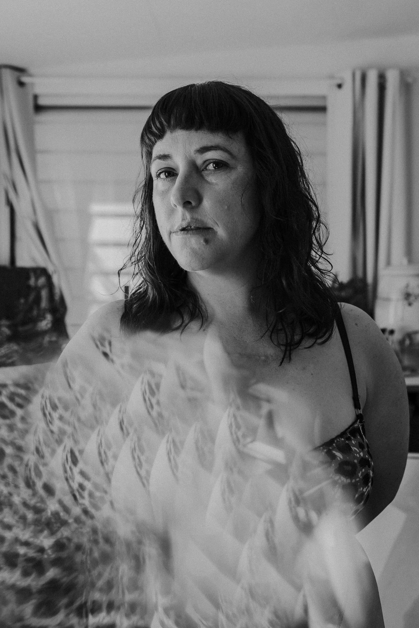 Black and white portrait of a woman with dark hair and bangs standing indoors with soft light filtering through curtains.