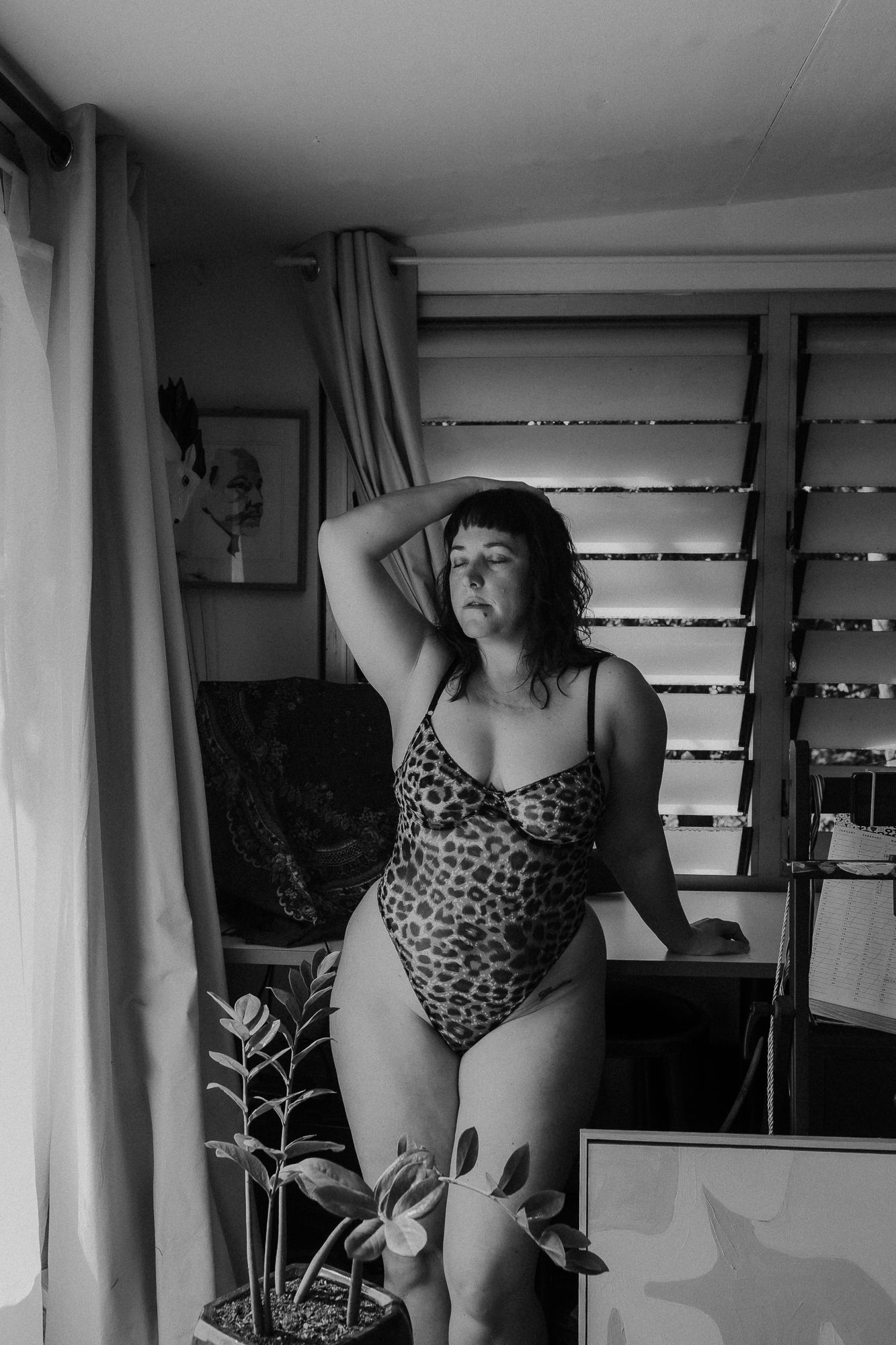 Woman in leopard print bodysuit poses confidently in a stylish room with plants and blinds in black and white photo.