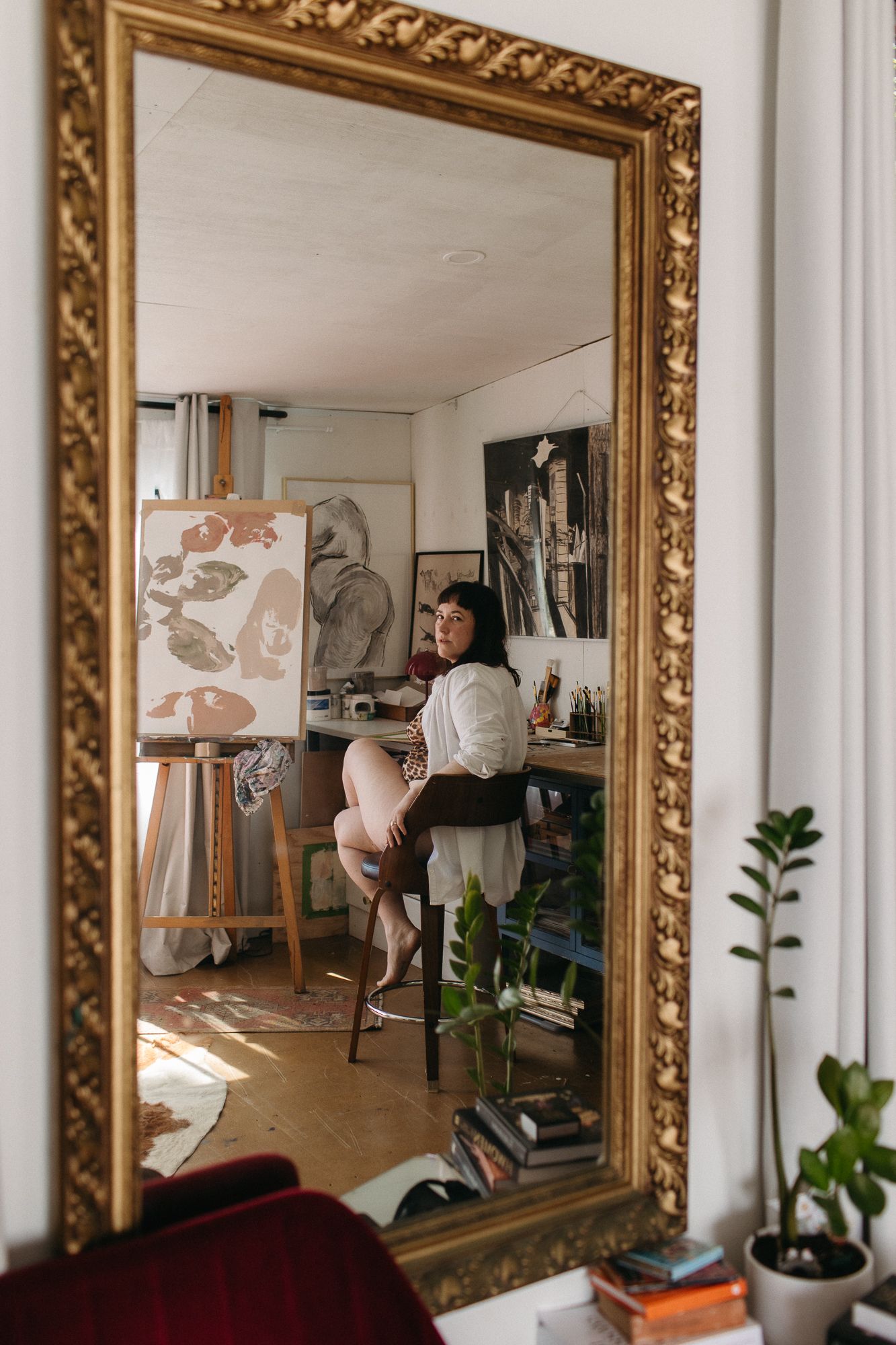 Artist reflected in ornate gold mirror in bright studio with easels, canvases, and plants.