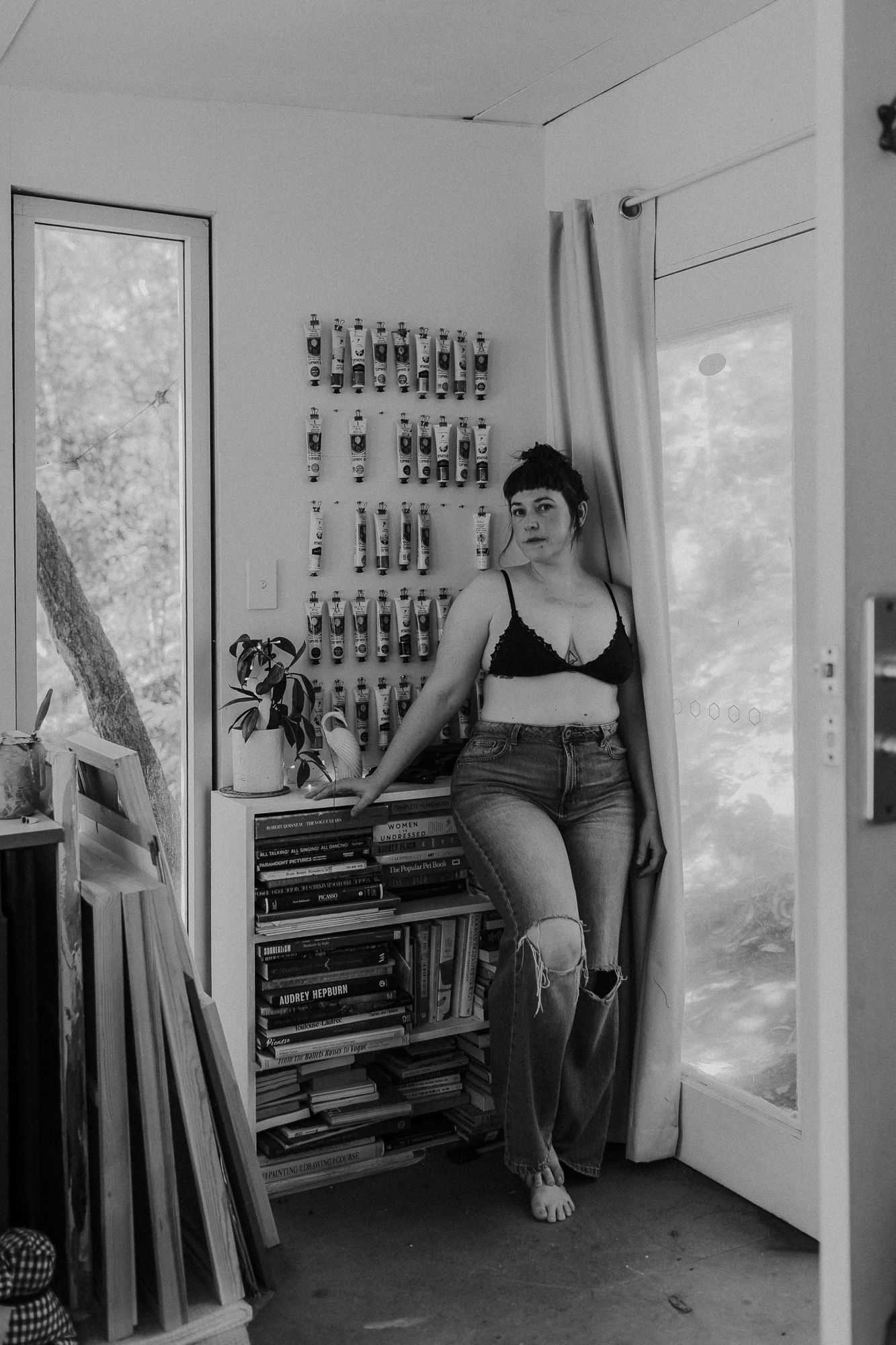 Black and white photo of a woman in a bralette and jeans leaning against a bookshelf in an art studio.