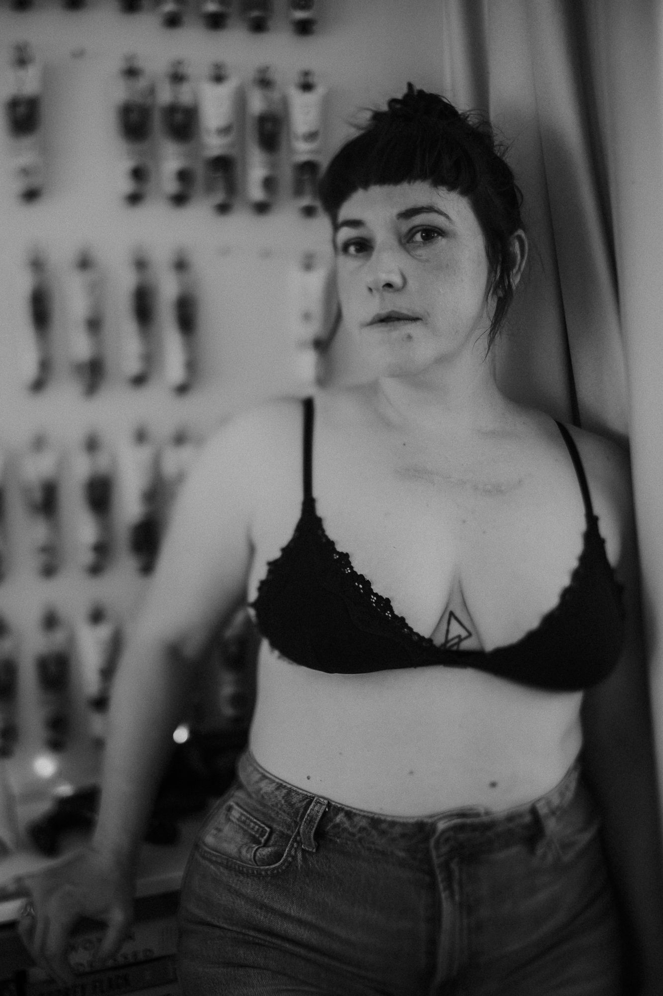 Black and white portrait of a woman in a black bralette and jeans with a chest tattoo leaning against a wall.