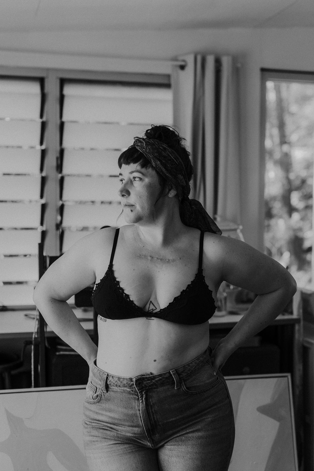 Black and white photo of a confident woman in a bralette and jeans posing indoors near a window.