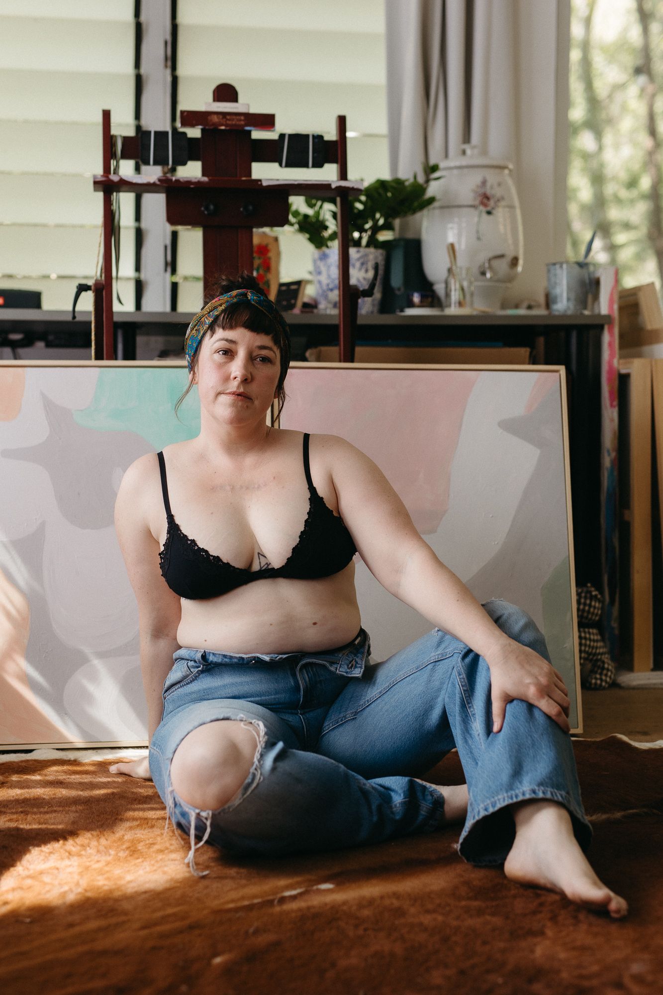 A plus-size woman in a black bralette and ripped jeans sits on the floor of an art studio with canvases behind her.