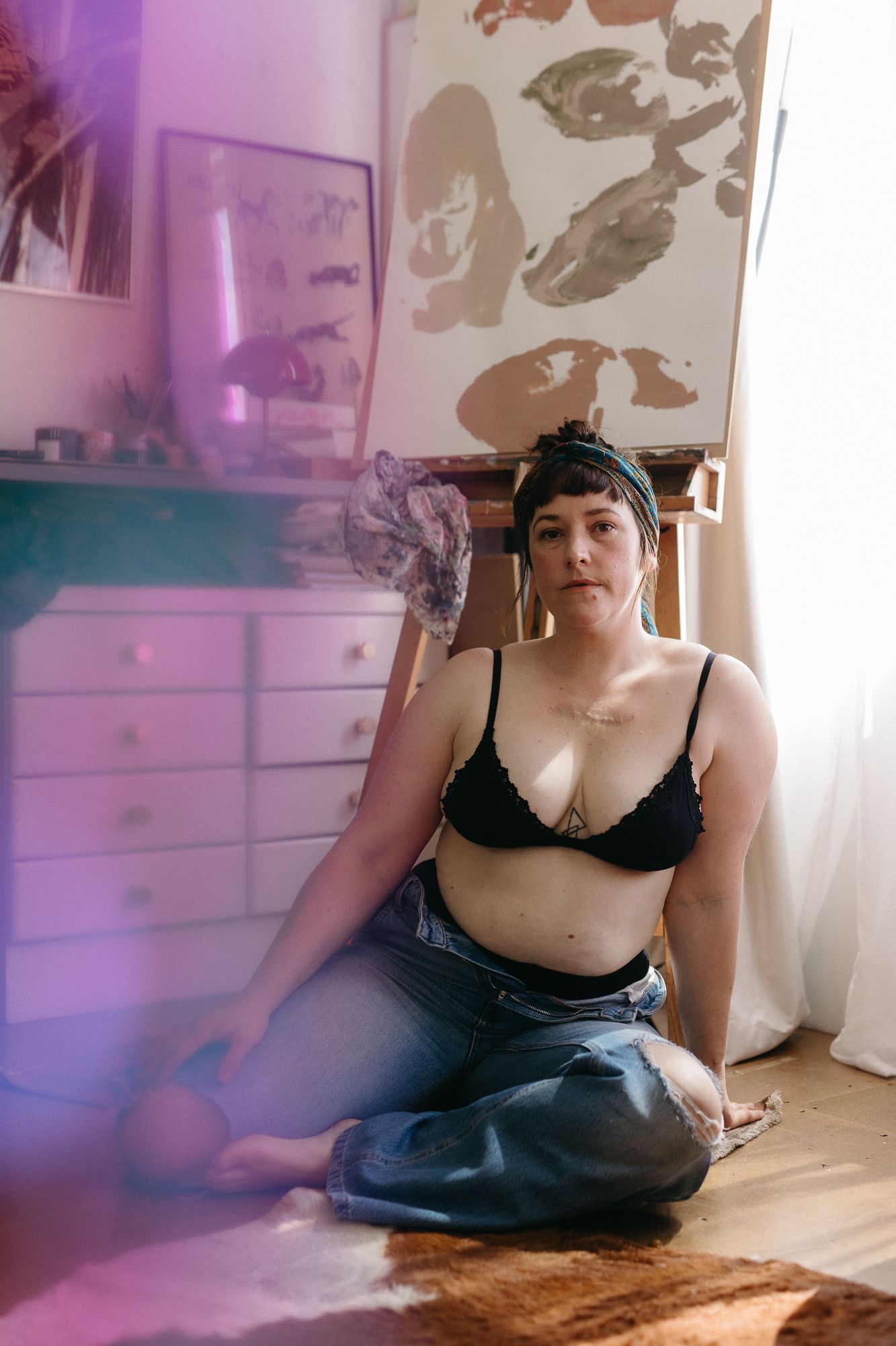 Woman in black bralette and jeans sitting on floor in bohemian bedroom with artwork and dresser.