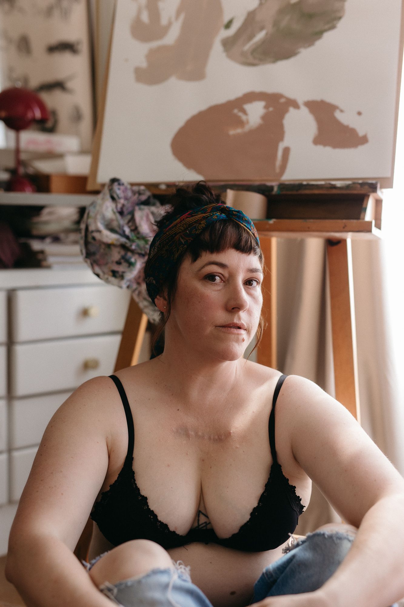 Artist in black lace bralette sitting in creative studio with easel and canvas painting in background.