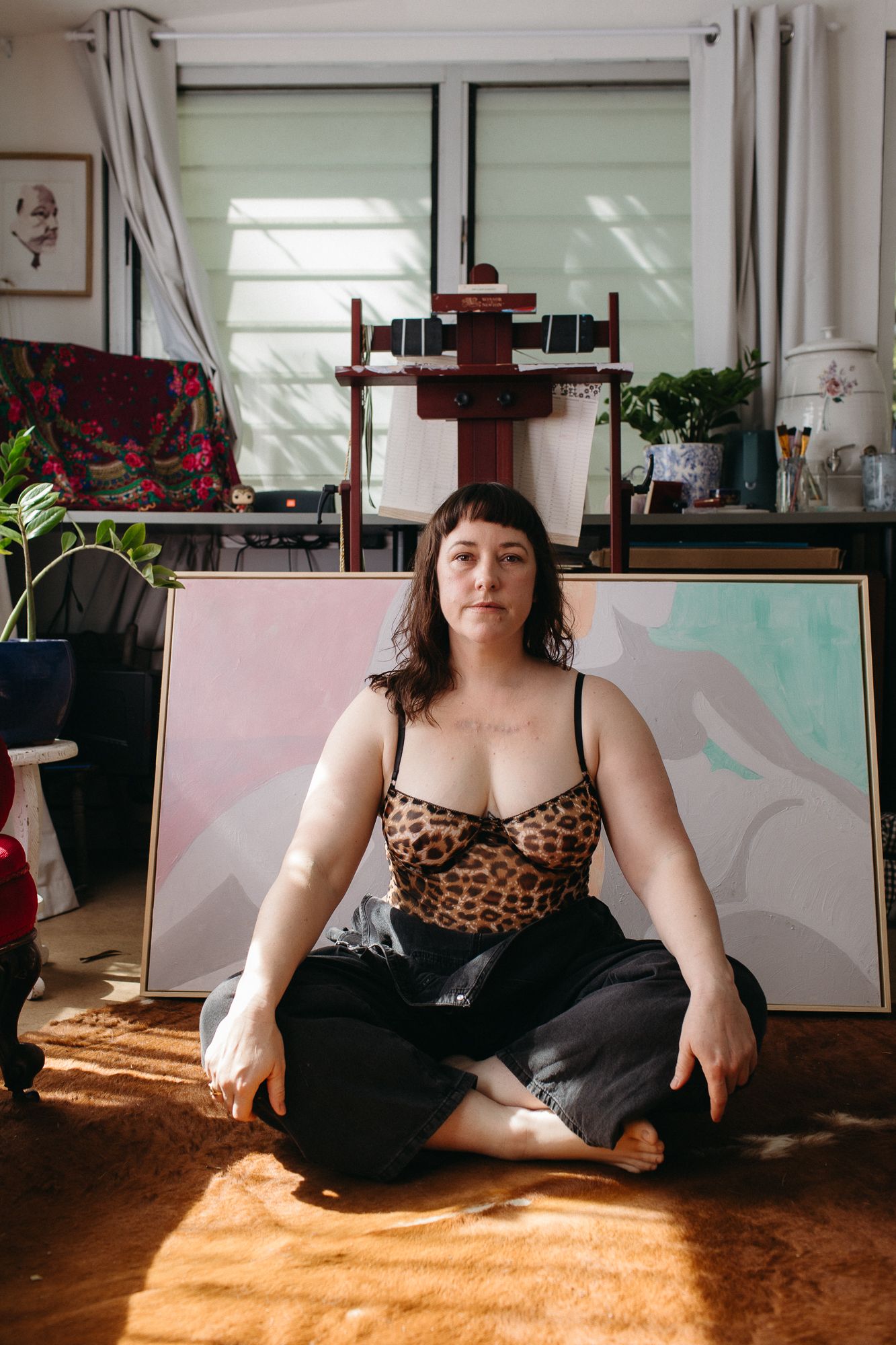 Woman in leopard print top sits cross-legged on floor of art studio surrounded by colorful paintings.