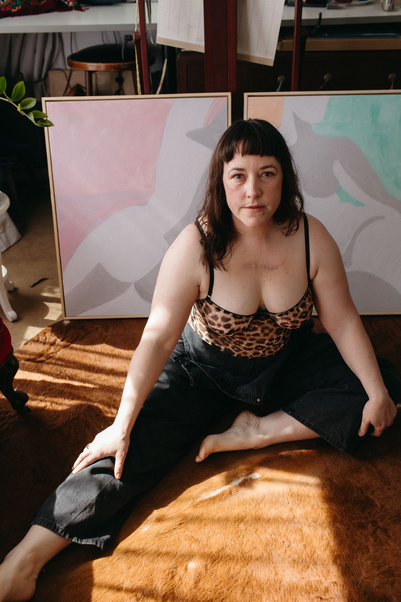 Woman in leopard print top sitting on floor in front of abstract pink and teal paintings in art studio.