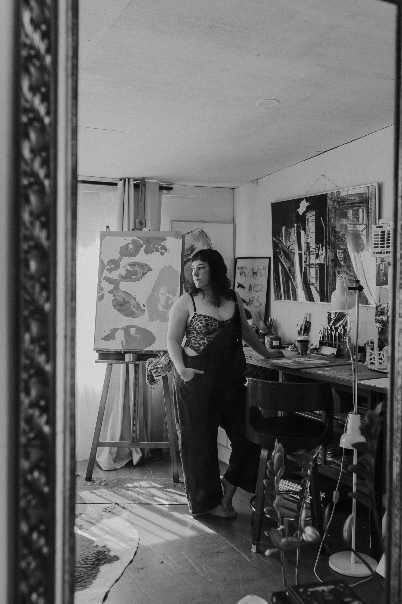 Black and white photo of a woman standing in an art studio with paintings on easel and walls behind her.