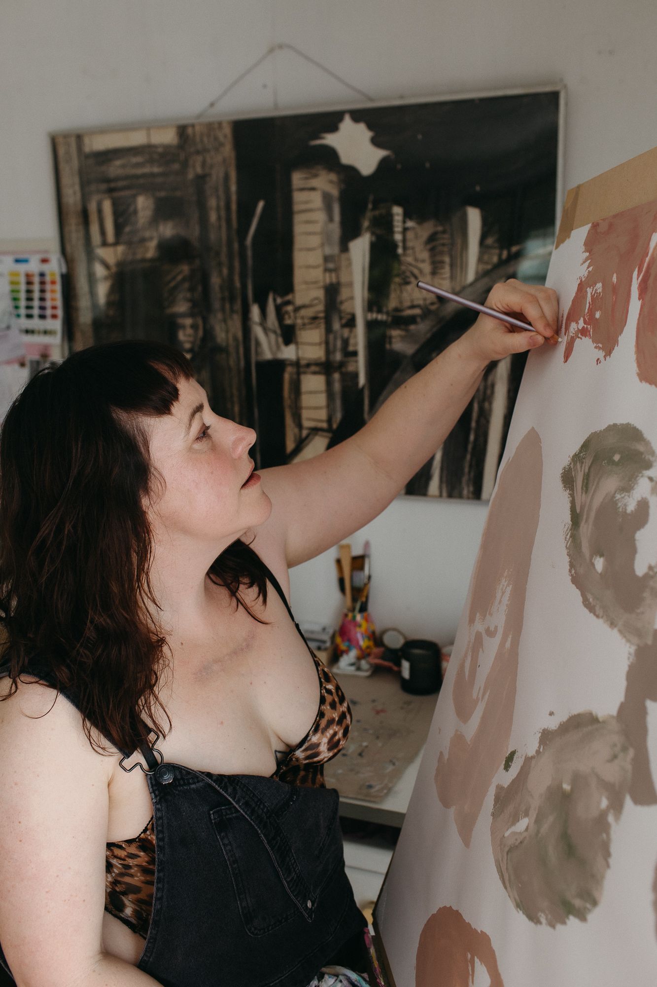 A woman in overalls painting on a canvas with a brush in a bright art studio.
