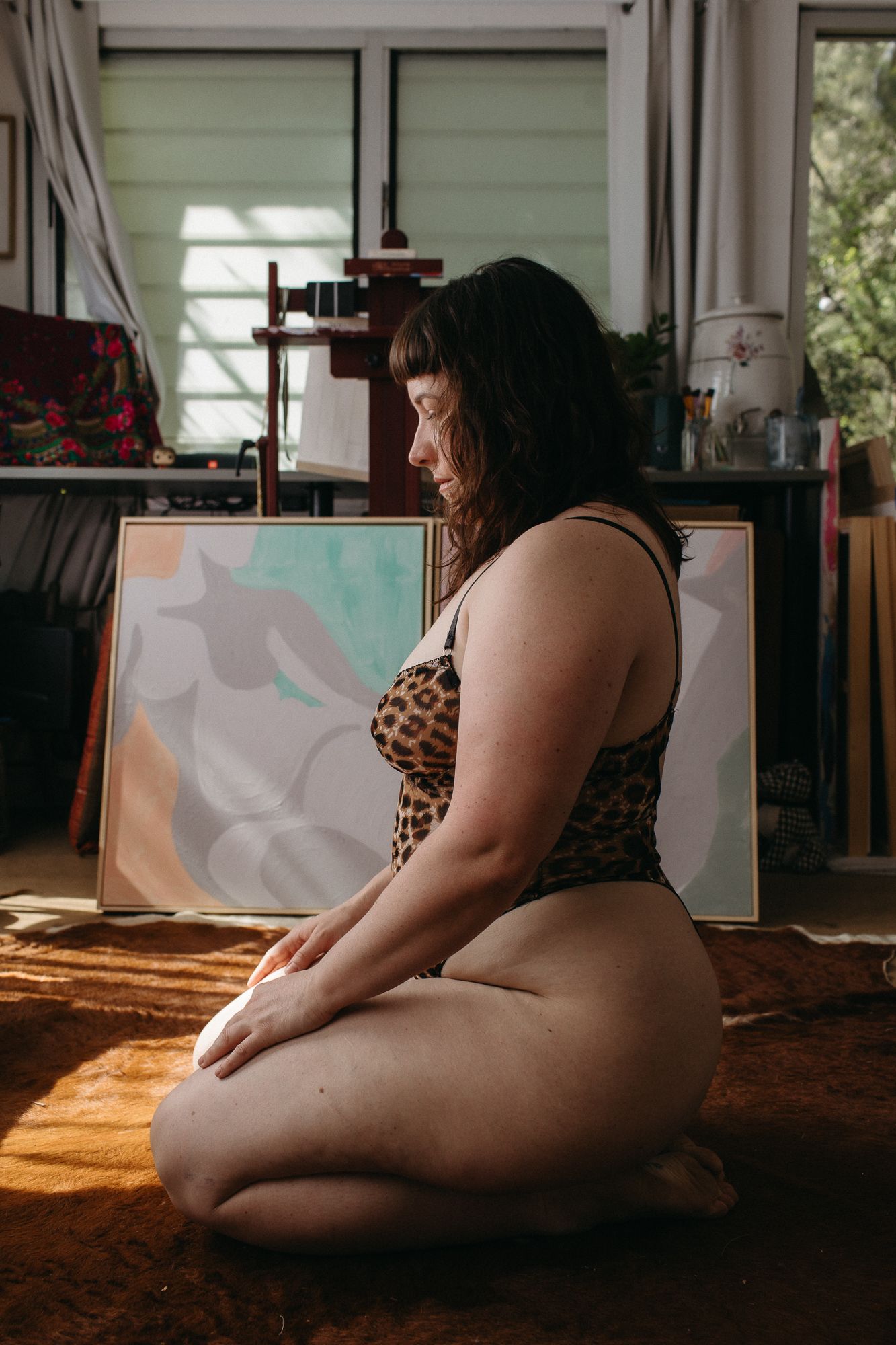 A plus-size woman in a leopard print bodysuit sits gracefully on a studio floor near colorful canvases.