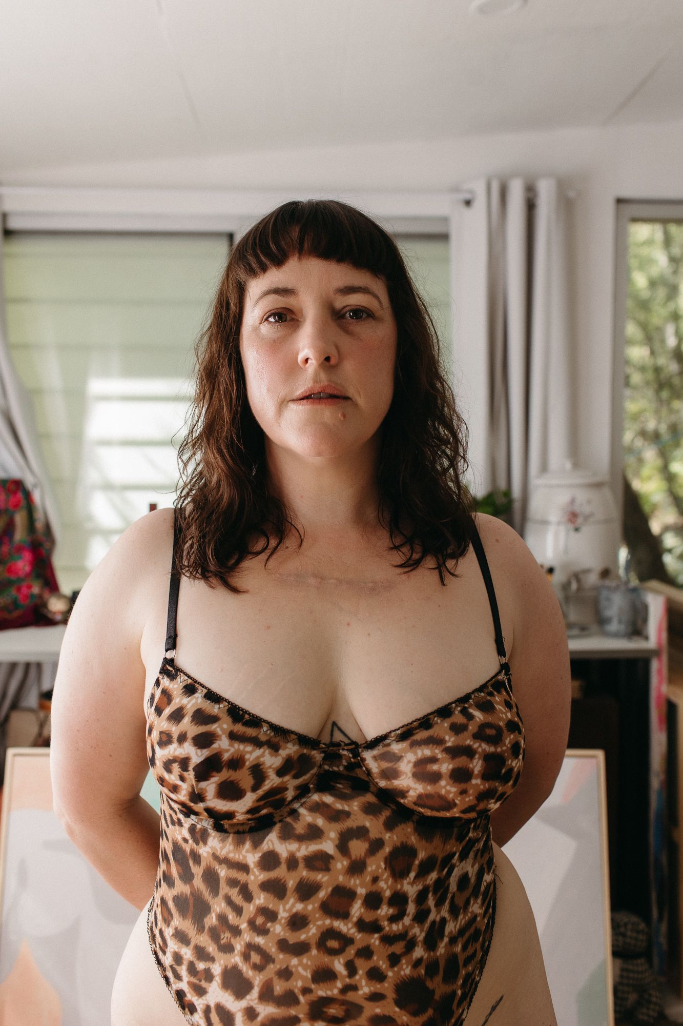 Woman in leopard print bodysuit posing indoors with neutral expression, dark hair, in a bright room.
