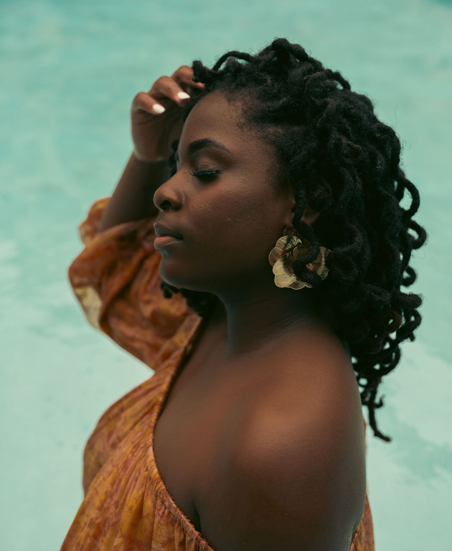 A Black woman with locs and gold floral earrings poses gracefully in an orange dress near a turquoise pool.