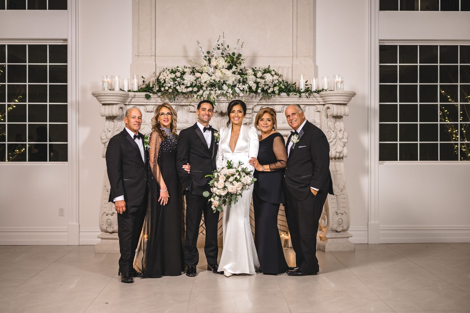 Formal wedding party portrait in an elegant white room with floral arrangements.