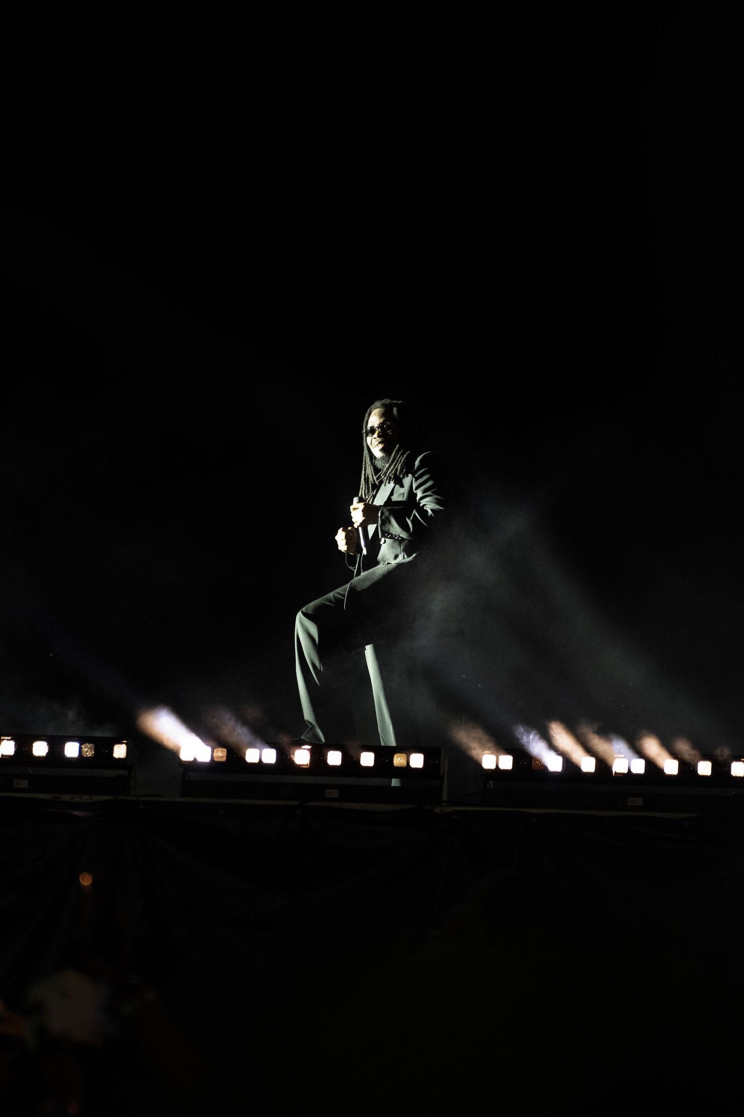 Performer stands illuminated on a dark stage with rows of bright lights and smoke effects during a concert.