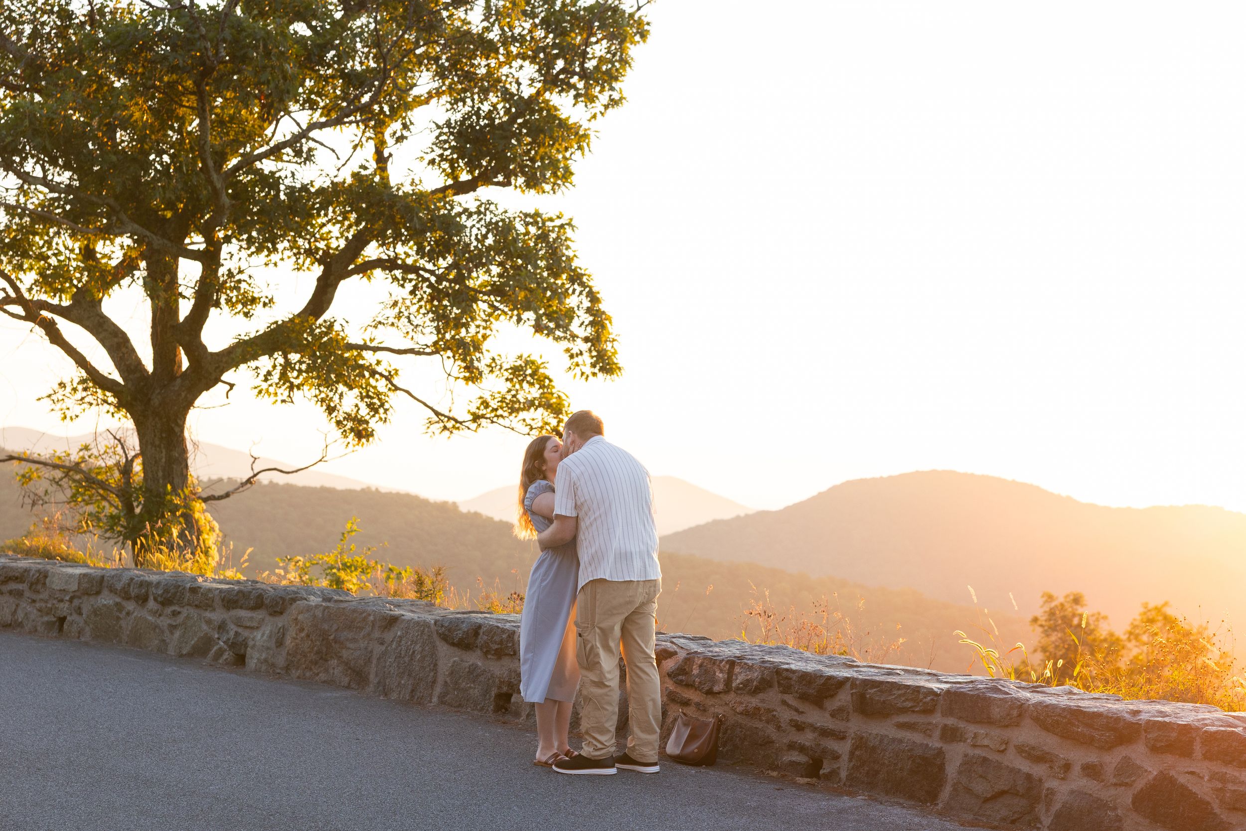 Couple sharing a romantic kiss at golden hour overlook with mountain views and a large oak tree.