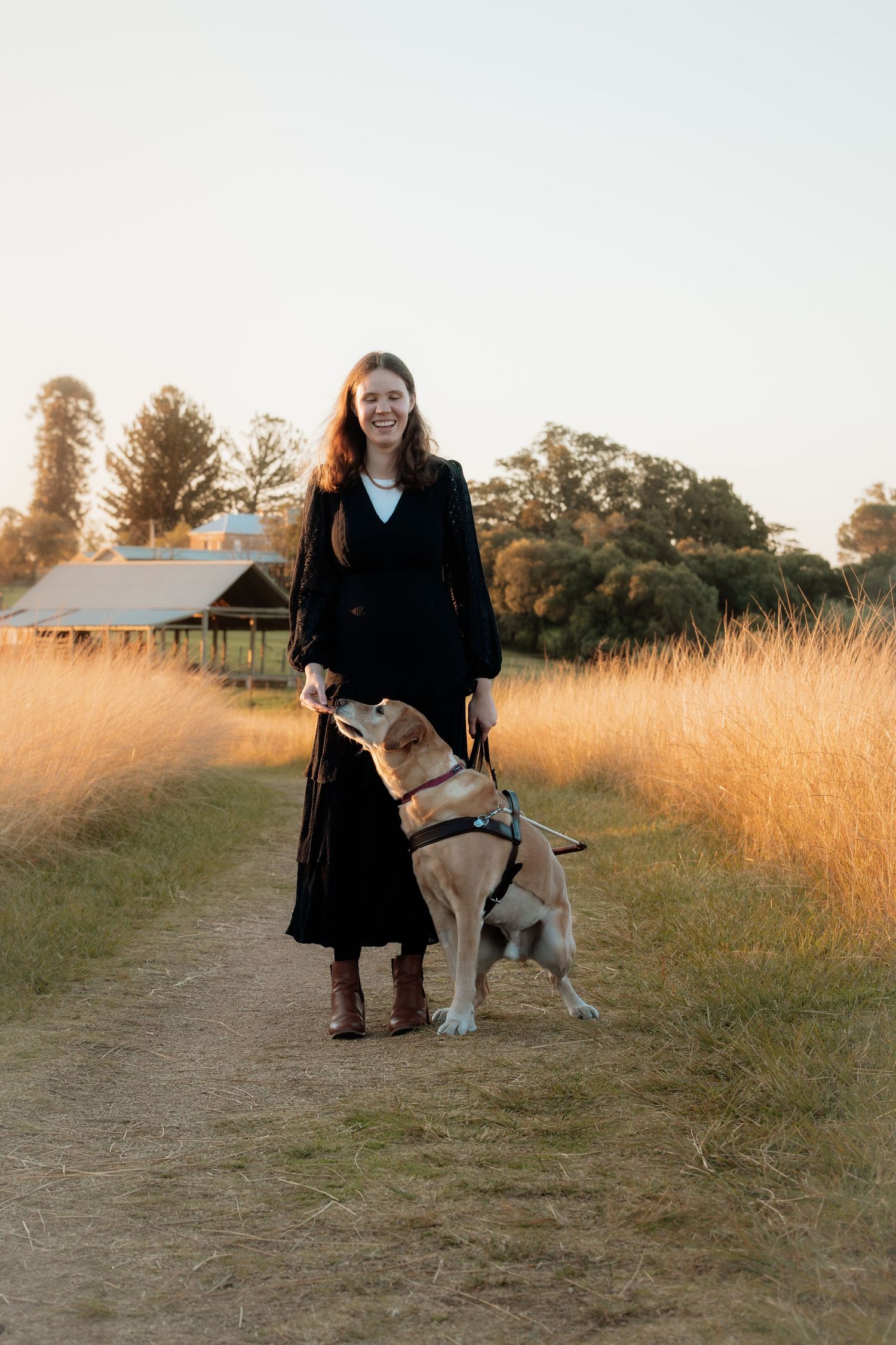 Woman in black dress and boots walks with a guide dog on a golden field path at sunset at Rouse Hill Regional Park.