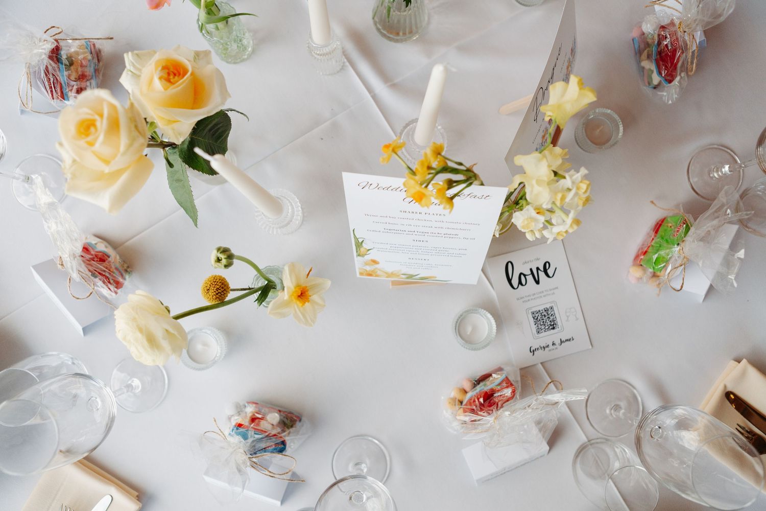 Wedding table setting with yellow roses, small bud vases, candles, wrapped favors, glassware, and a QR code sign on a white tablecloth.
