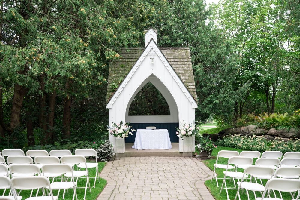 Outdoor garden wedding ceremony site with a white gothic-style gazebo, floral arrangements, and white folding chairs along a brick path.