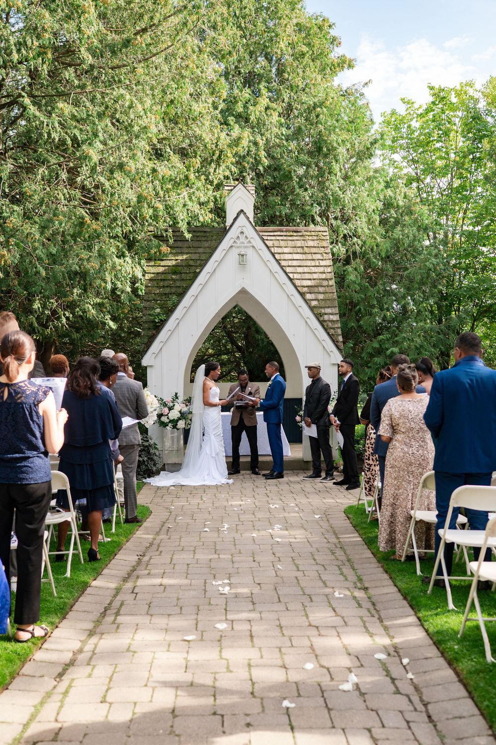 Bride and groom exchange vows with an officiant under a white gothic chapel archway as guests watch from a brick path.