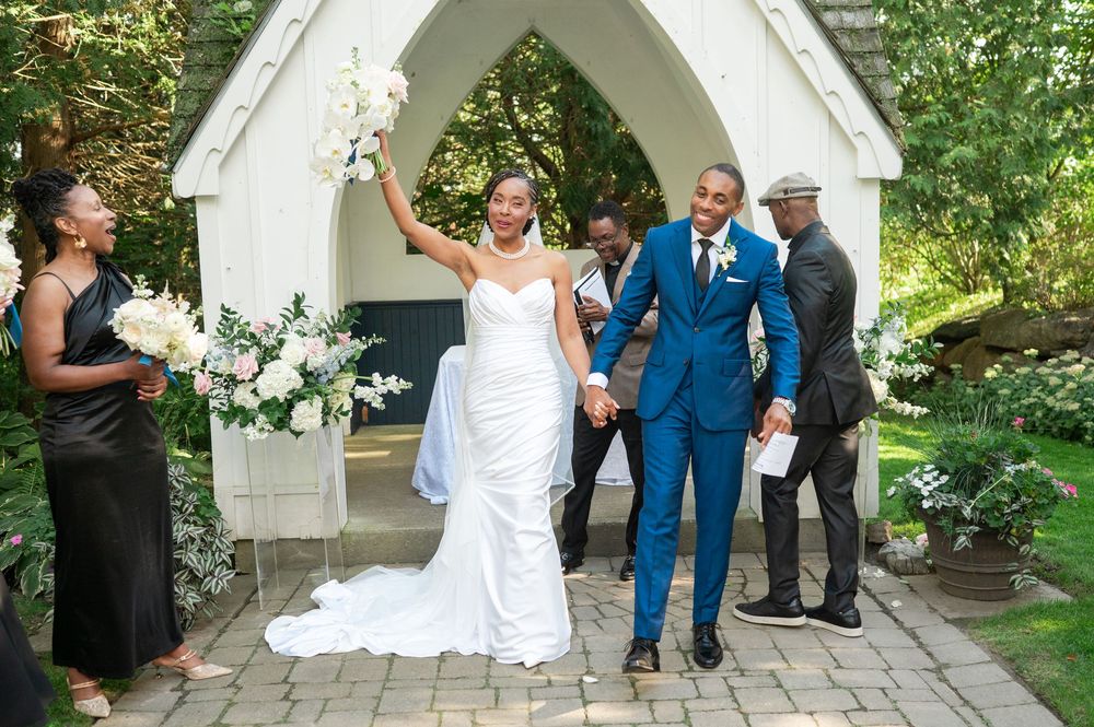 Bride in a white gown and groom in a bright blue suit hold hands outside a white stone garden chapel, surrounded by guests and flowers.
