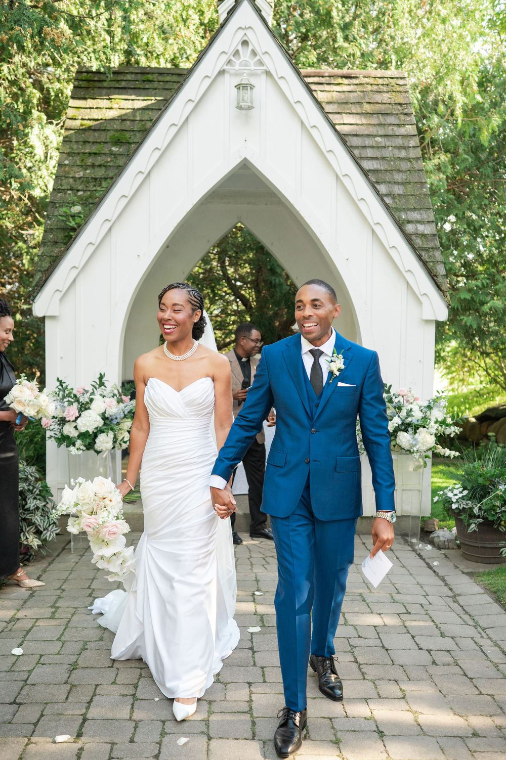 Smiling newlyweds hold hands as they walk out of a white garden chapel, the bride in a strapless gown and the groom in a blue suit.