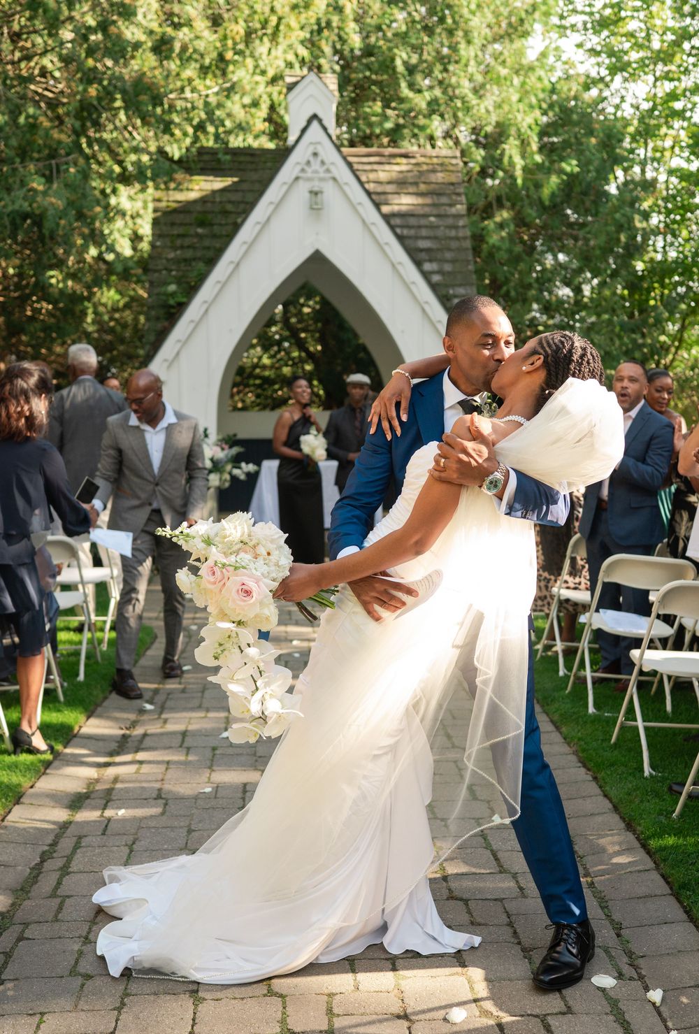 Groom in a blue suit dips and kisses his bride in a flowing white veil holding a bouquet during an outdoor wedding ceremony with guests.