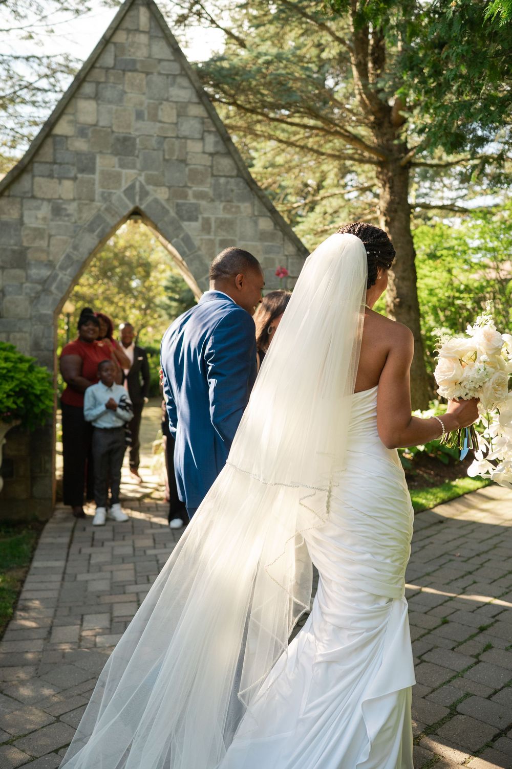 Bride in a white gown and long veil walks with her groom in a blue suit along a brick path toward a stone arch at an outdoor wedding.