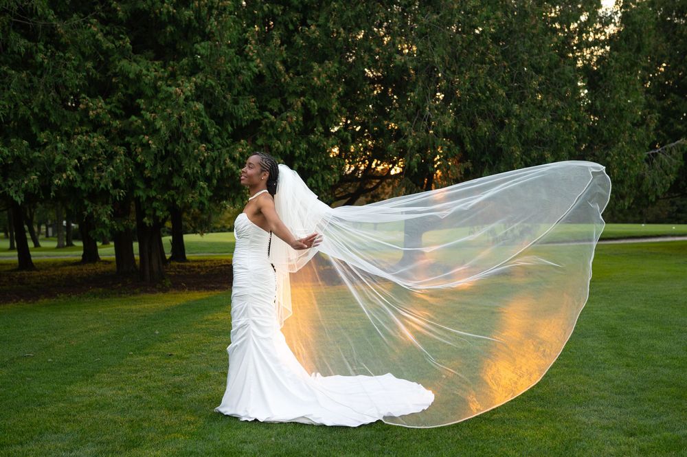 Bride in a white strapless wedding gown holds her long sheer veil as it billows in the breeze on a green lawn lit by golden sunset light.