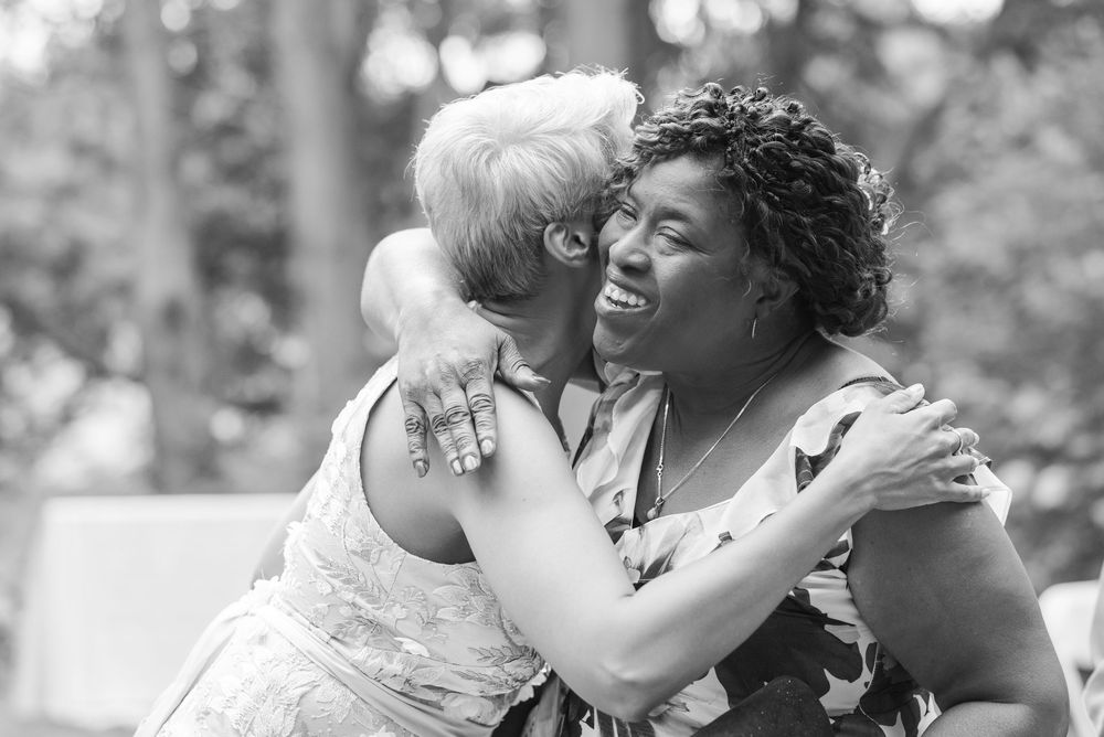 Two women share a joyful hug outdoors in a black and white photo, one laughing with closed eyes, surrounded by blurred trees.