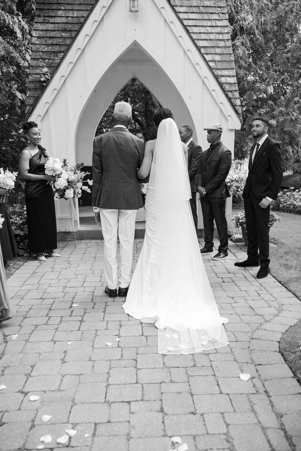Black and white photo of a bride in a long veil walking down a stone path toward a small chapel, escorted by a man as guests watch.