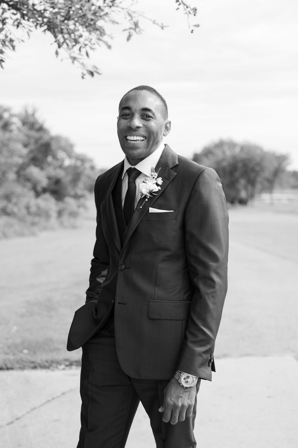 Smiling groom in a dark suit with a floral boutonniere and pocket square poses outdoors in a black and white wedding portrait.