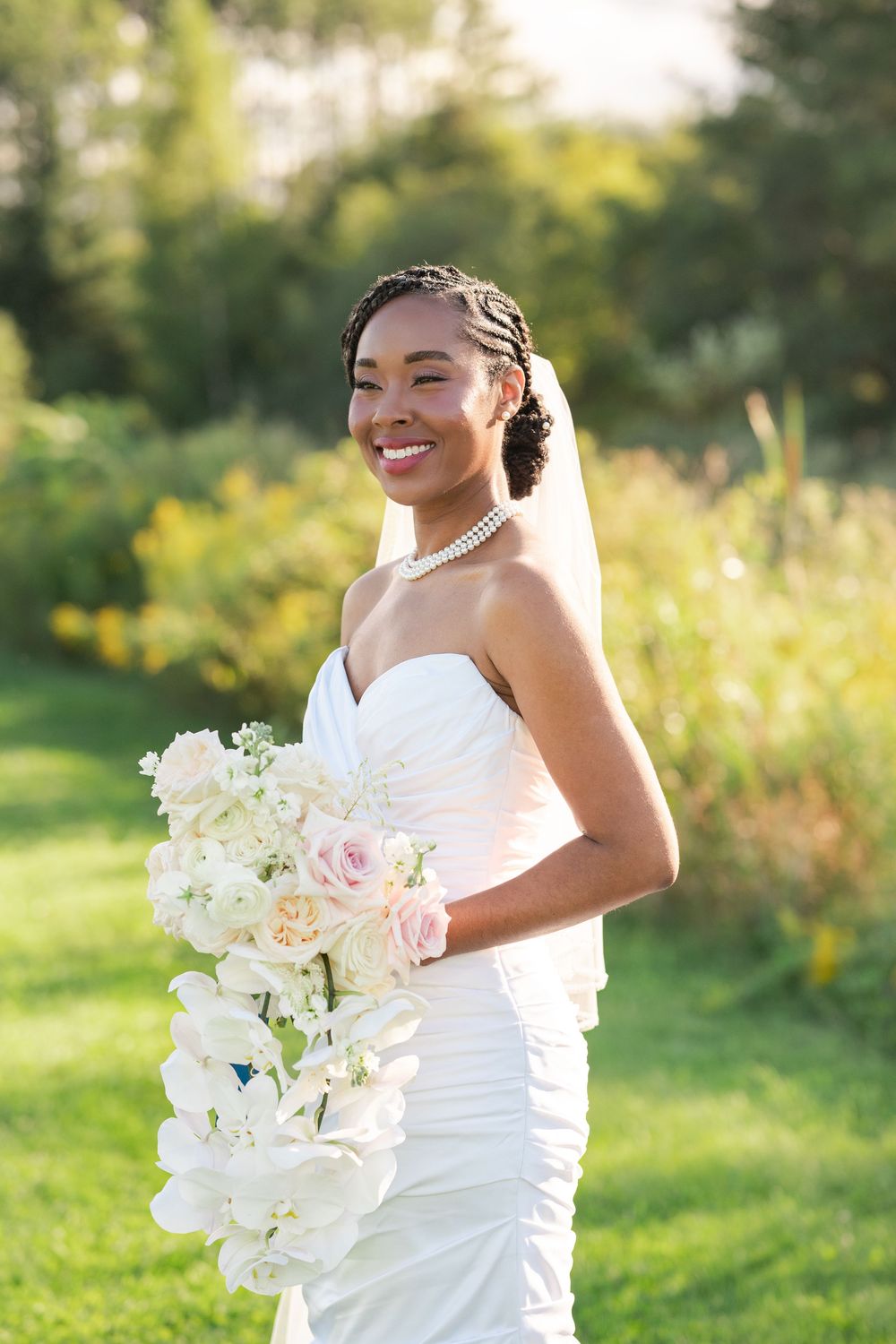 Smiling bride in a white strapless wedding gown and veil holds a cascading bouquet of white and blush flowers in a sunlit green meadow.