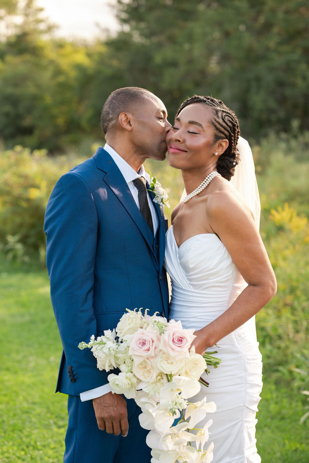 Groom in a blue suit kisses his smiling bride on the cheek while she holds a white rose and orchid bouquet outdoors.