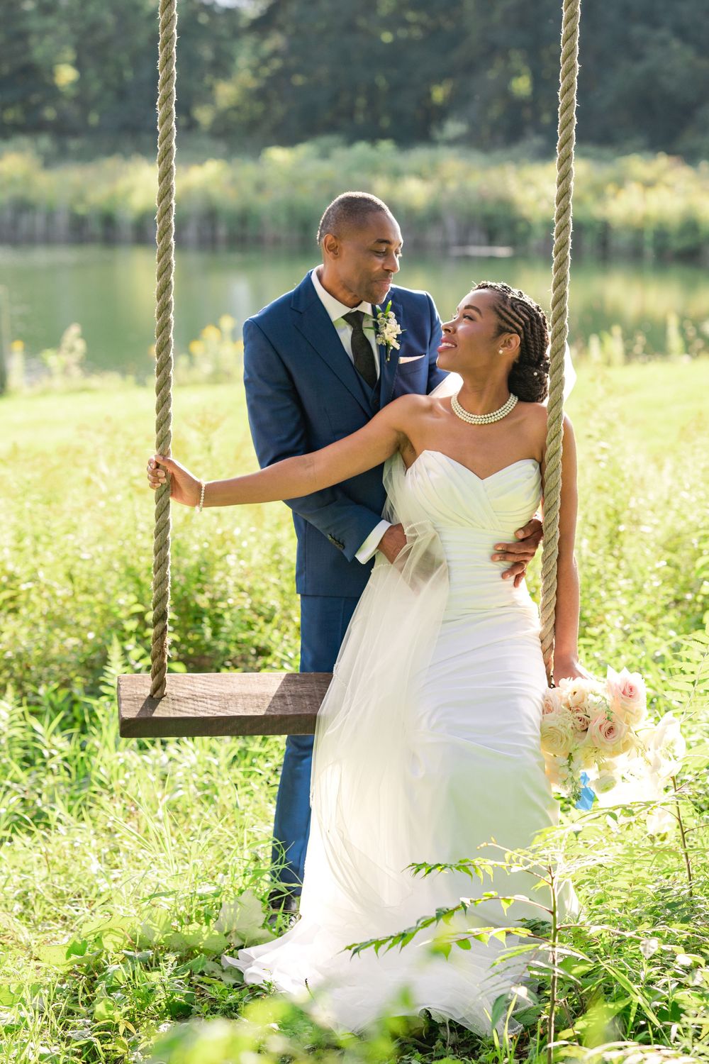 Bride in a white strapless gown holding a bouquet and smiling at her groom in a navy suit beside a rope swing in a grassy field by a lake.