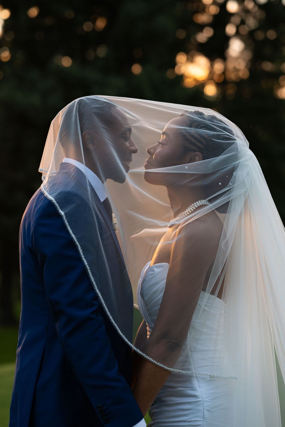 Bride and groom share a romantic moment beneath a sheer veil with glowing golden sunset light behind them.